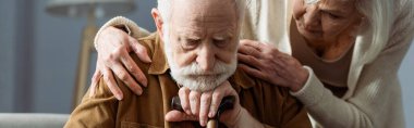 Yaşlı adamın panoramik görüntüsü, bunama hastası, bastonla oturuyor, karısı ona bakıyor ve omuzlarına dokunuyor.