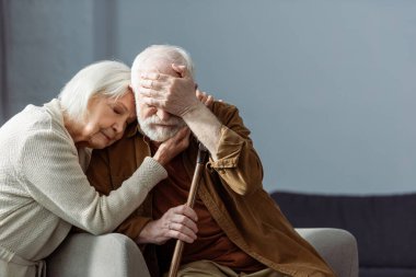 senior man, sick on dementia, covering eyes with hand while wife embracing him with closed eyes clipart