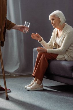 partial view of senior man giving glass of water to wife feeling bad and sitting on sofa clipart