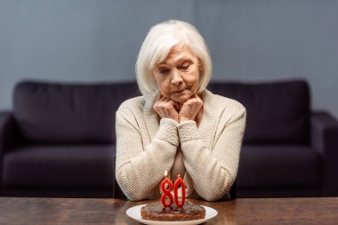 thoughtful, sad senior woman sitting near birthday cake with number eighty and burning candles alone clipart