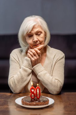 lonely senior woman looking at birthday cake with number eighty and burning candles  clipart
