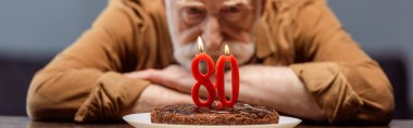selective focus of lonely senior man looking at birthday cake with number eighty, horizontal image clipart