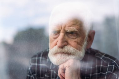 grieving senior man looking away through window glass while holding hand near chin clipart