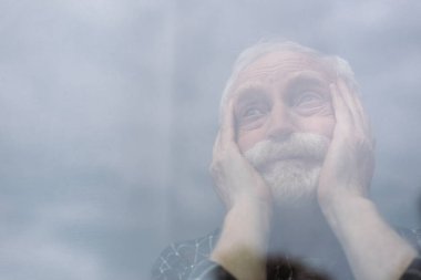 selective focus of senior, lonely man touching face and looking away through window glass clipart
