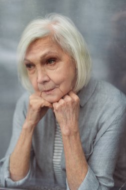 sad senior woman looking away through window glass clipart