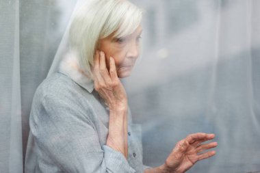 depressed senior woman touching face and window glass while looking away clipart