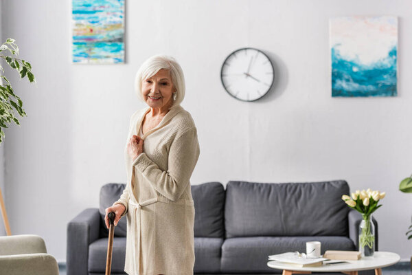 happy senior woman looking at camera while standing with walking stick at home
