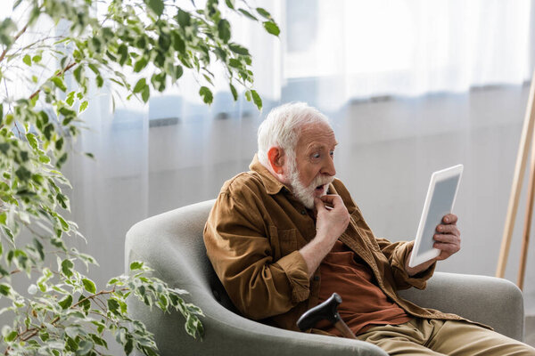 потрясенный пожилой человек с помощью цифрового планшета, сидя в кресле
