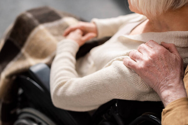 cropped view of man touching shoulder of handicapped, diseased wife sitting in wheelchair