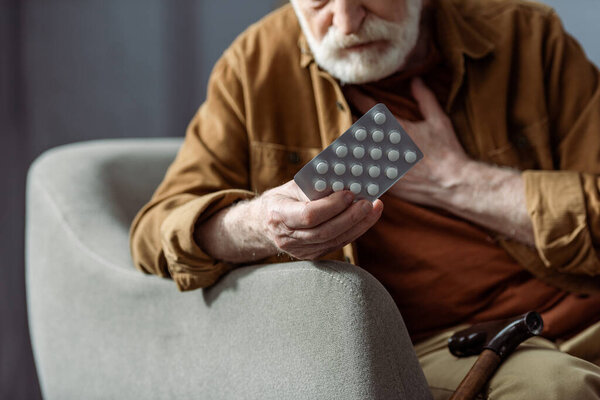 selective focus of senior man touching chest and holding pills while feeling bad