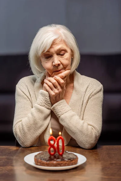 lonely senior woman looking at birthday cake with number eighty and burning candles