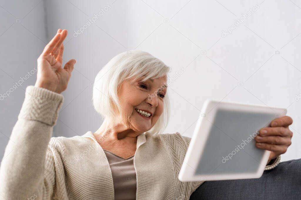 cheerful senior woman sitting with raised hand while using digital tablet