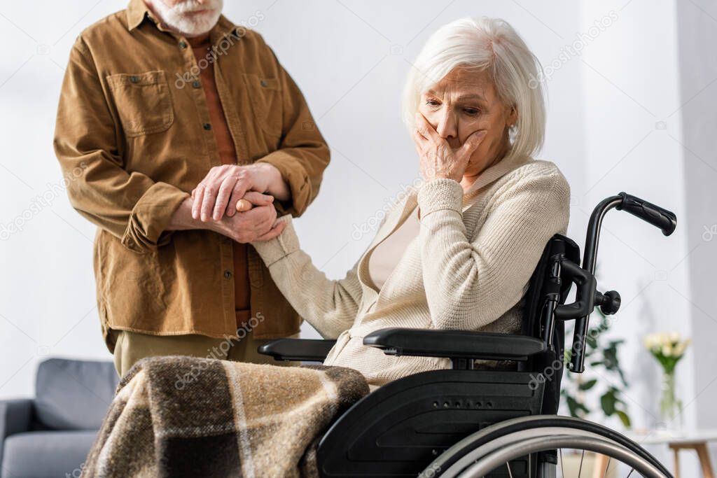 cropped view of man holding hand of disabled wife, sick on dementia, holding hand on face while sitting in wheelchair