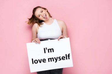 cheerful overweight girl holding placard with i love myself inscription and sticking out tongue on pink  clipart