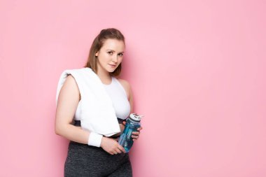 Omzunda havluyla spor şişesi tutan ve pembe kameraya bakan şişman güzel bir kız.
