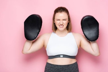 angry overweight girl in boxing pads grimacing while looking at camera on pink clipart