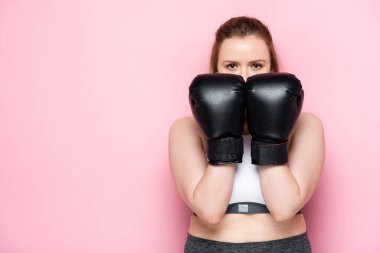 confident overweight girl obscuring face with boxing gloves while looking at camera on pink clipart