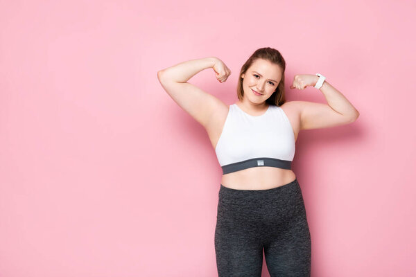 cheerful overweight girl in sportswear demonstrating biceps on pink