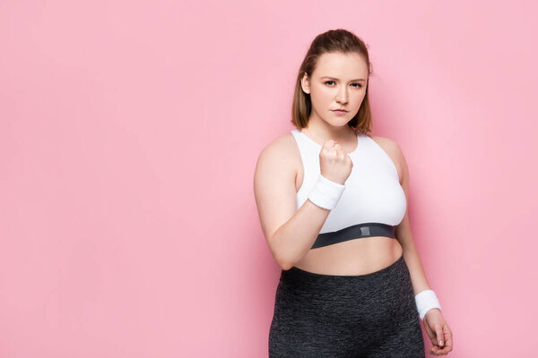 сердитая толстая девушка показывает сжатый кулак и смотрит в камеру на розовый

