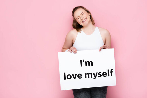 cheerful overweight girl with closed eyes holding placard with I love myself inscription on pink 