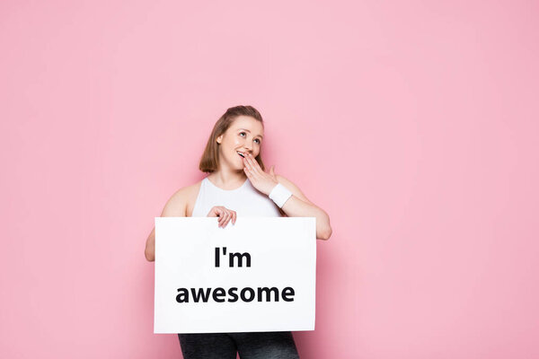 flirty overweight girl holding placard with I am awesome inscription on pink