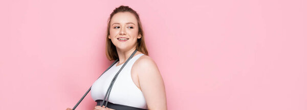 horizontal image of smiling overweight girl with jumping rope around neck looking away on pink