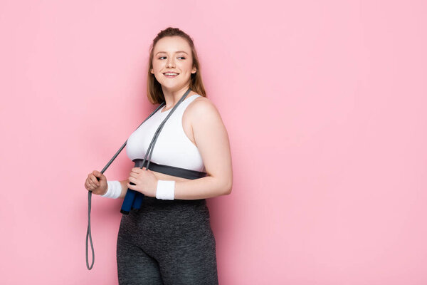 smiling overweight girl with jumping rope around neck looking away on pink