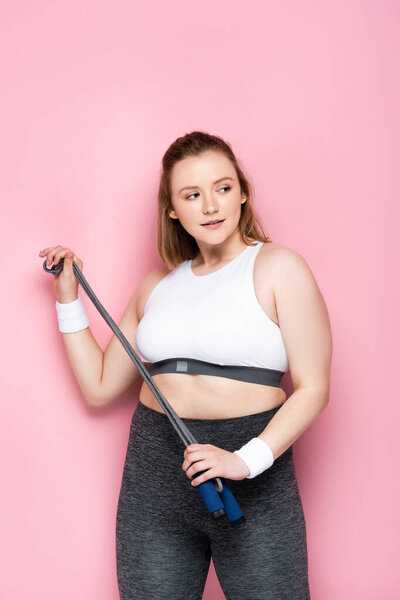привлекательная толстая девушка держит скакалку, отводя взгляд на розовый
