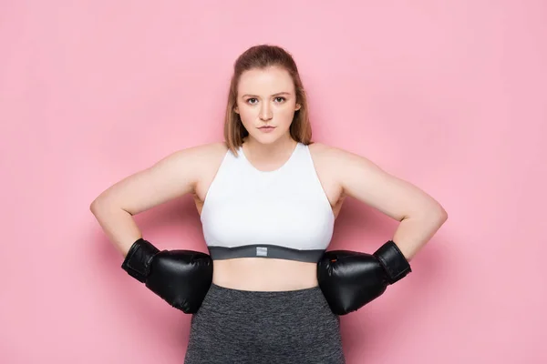 confident size plus girl in boxing gloves holding hands on hips while looking at camera on pink