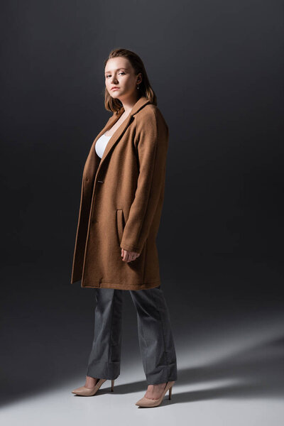 красивая, трогательная девушка с лишним весом, смотрящая на камеру, позируя в одиночестве на сером фоне
