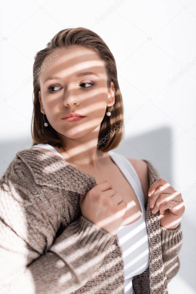 confident, attractive plus size girl touching sweater while looking away on white with sunlight and shadows