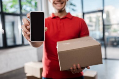 cropped view of smiling delivery man holding smartphone with blank screen and carton box  clipart