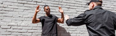 panoramic orientation of policeman in cap detaining african american man with raised hands, racism concept  clipart