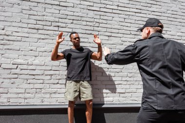 policeman in cap detaining african american man with raised hands, racism concept  clipart