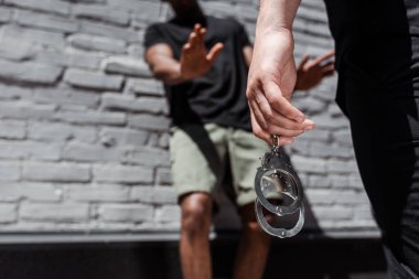 cropped view of police officer holding handcuffs while detaining african american man gesturing near brick wall, racism concept  clipart