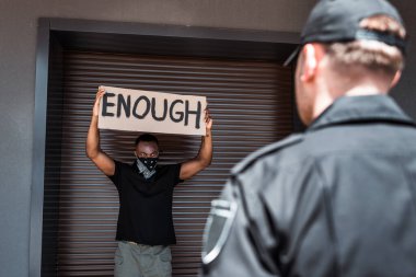 selective focus of african american man with scarf on face holding placard with enough lettering near policeman on street, racism concept  clipart