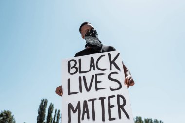Yüzünde fular olan Afro-Amerikan bir adamın düşük açılı görüntüsü. Elinde siyah yaşam yazılı bir pankart var. Gökyüzüne karşı yazılmış.