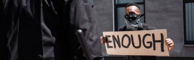 selective focus of african american man with scarf on face holding placard with enough lettering near policeman with baton on street, racism concept  clipart