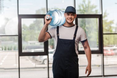 cheerful delivery man in cap and uniform holding gallon of water in bottle  clipart