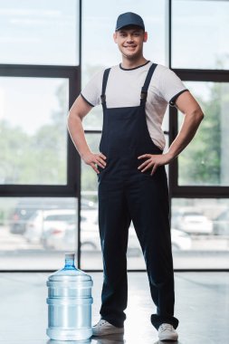 cheerful delivery man in uniform standing with hands on hips near bottled water  clipart