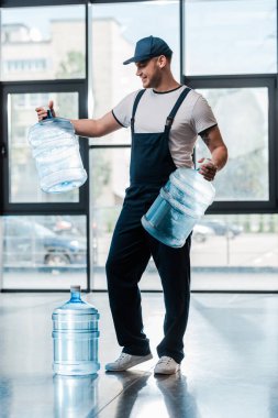 cheerful delivery man in uniform looking at empty bottles near gallon of water  clipart