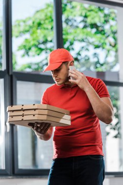 Kapüşonlu teslimatçı akıllı telefondan konuşuyor ve elinde pizza kutuları var. 