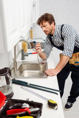 Selective focus of plumber in overalls holding metal pipe near sink in kitchen  clipart