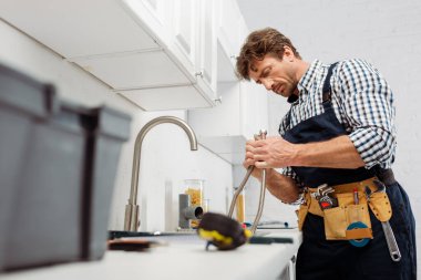 Selective focus of plumber holding metal pipe near kitchen faucet and tools on worktop  clipart