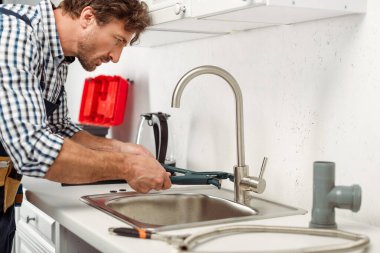 Side view of handsome repairman using pipe wrench while fixing kitchen faucet  clipart