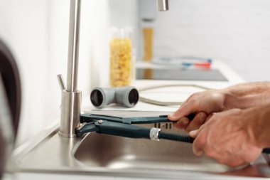Cropped view of plumber fixing kitchen faucet with pipe wrench near pipes on worktop  clipart