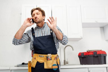 Handsome plumber in overalls and tool belt talking on smartphone in kitchen  clipart