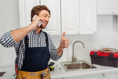 Gülümseyen tesisatçı jest yapıyor ve mutfak lavabosunun yanında akıllı telefondan konuşuyor. 