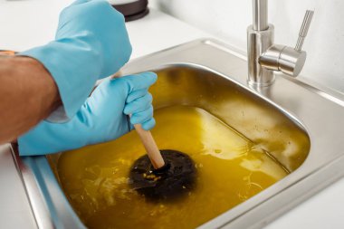 Mutfak lavabosunun tıkanmasını temizlerken elinde pompa tutan lastik eldivenli tesisatçı görüntüsü. 