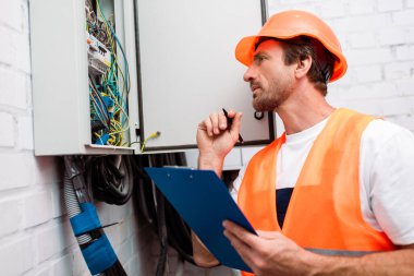 Selective focus of handsome electrician in safety vest and hardhat holding pen and clipboard while looking at electric panel clipart